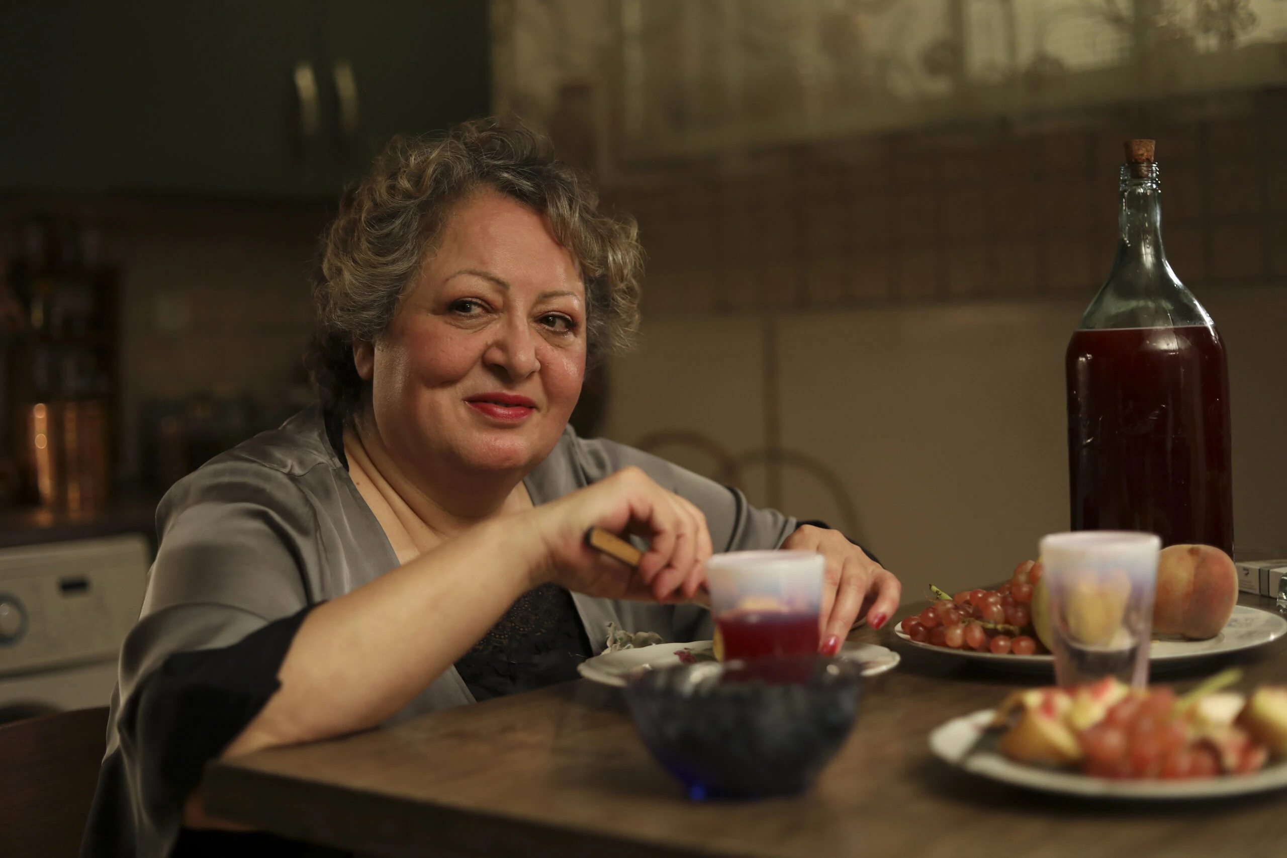 Una donna anziana sorride seduta a tavola con bicchieri e piatti di frutta.