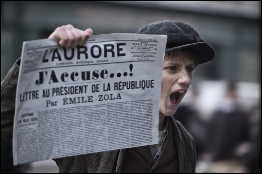 Un giovane strillone mostra una copia del giornale francese L'Aurore con il celebre articolo «J'accuse…!» di Émile Zola, in una scena del film L'ufficiale e la spia (2019)